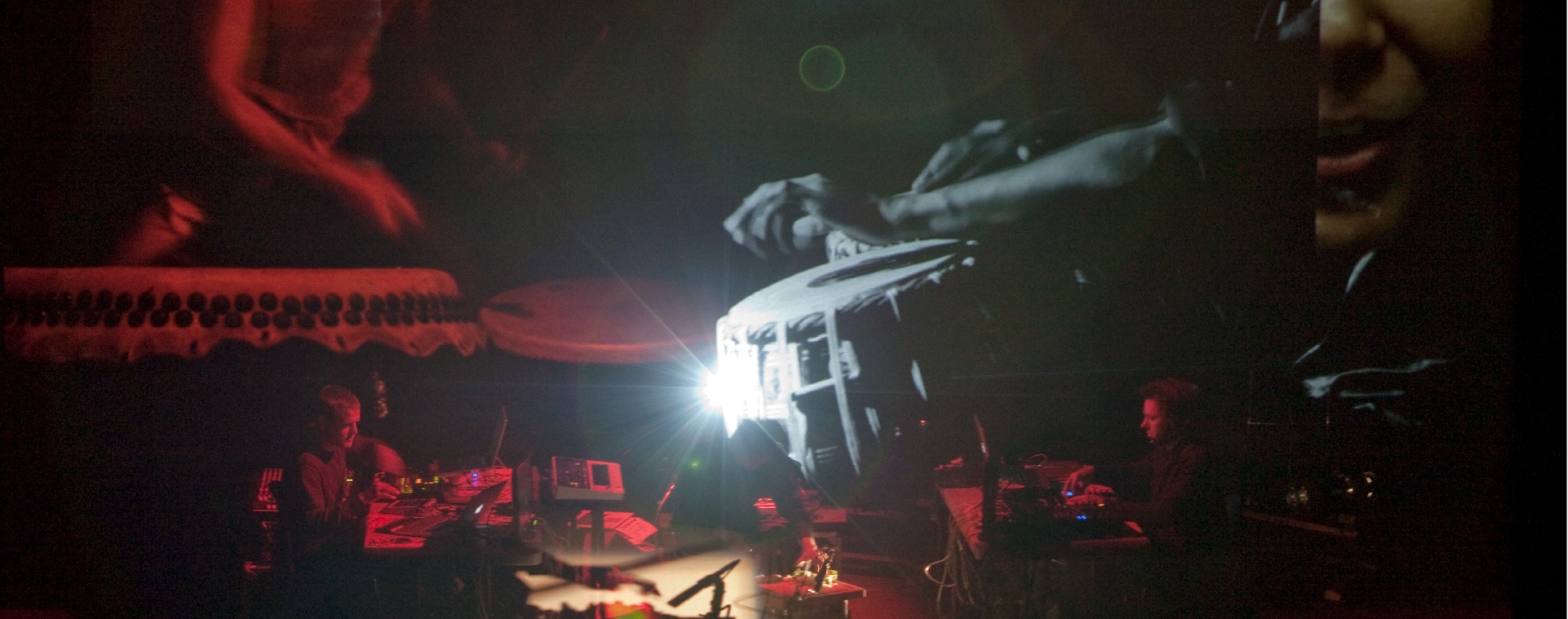 Silhouetted artists on stage use computers and technology to control projections on a large screen above them showing people playing hand drums in red and grey light.