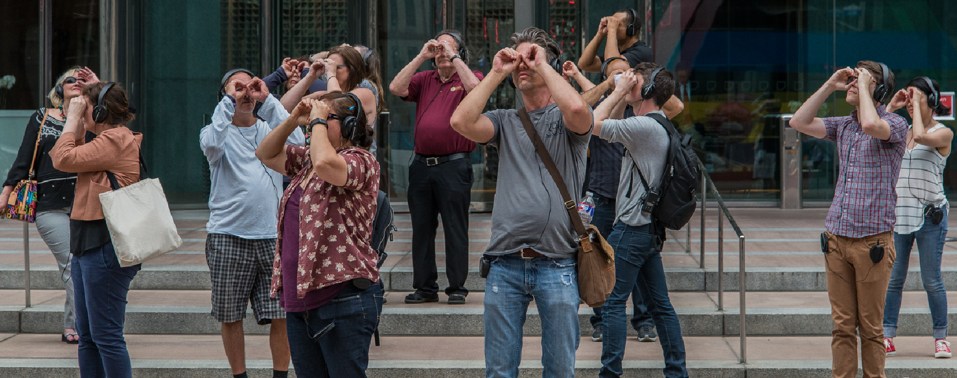 A group of people facing different directions stand on the steps to the entrance to an office building with headphones on, looking through their hands like binoculars.