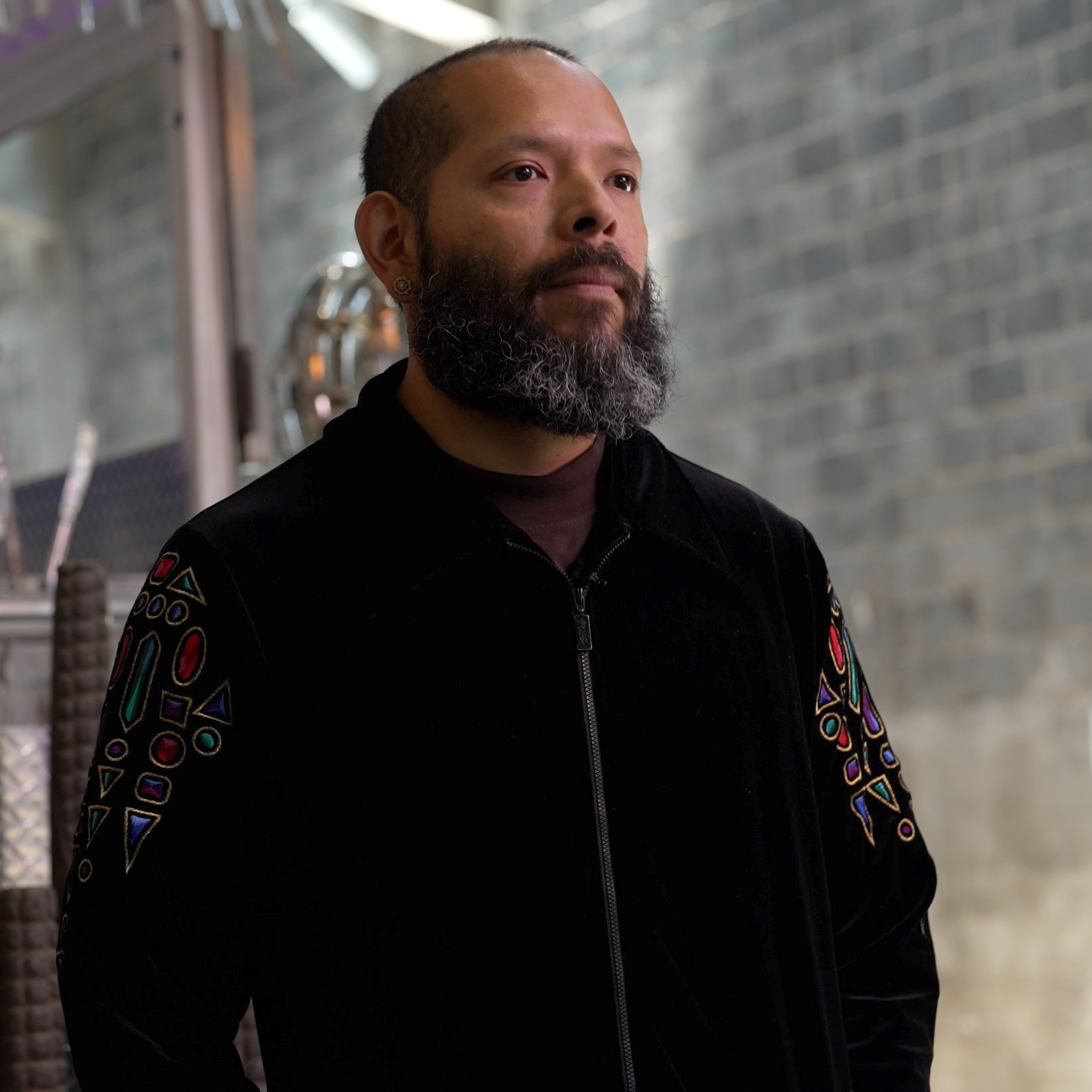 an El Salvadorian man with a mid-length beard and hair cut close to his head looks away from the camera. he is wearing a black jacket that looks to be made of a reflective material such as satin. there is decorative embroidery or patches near the shoulders.