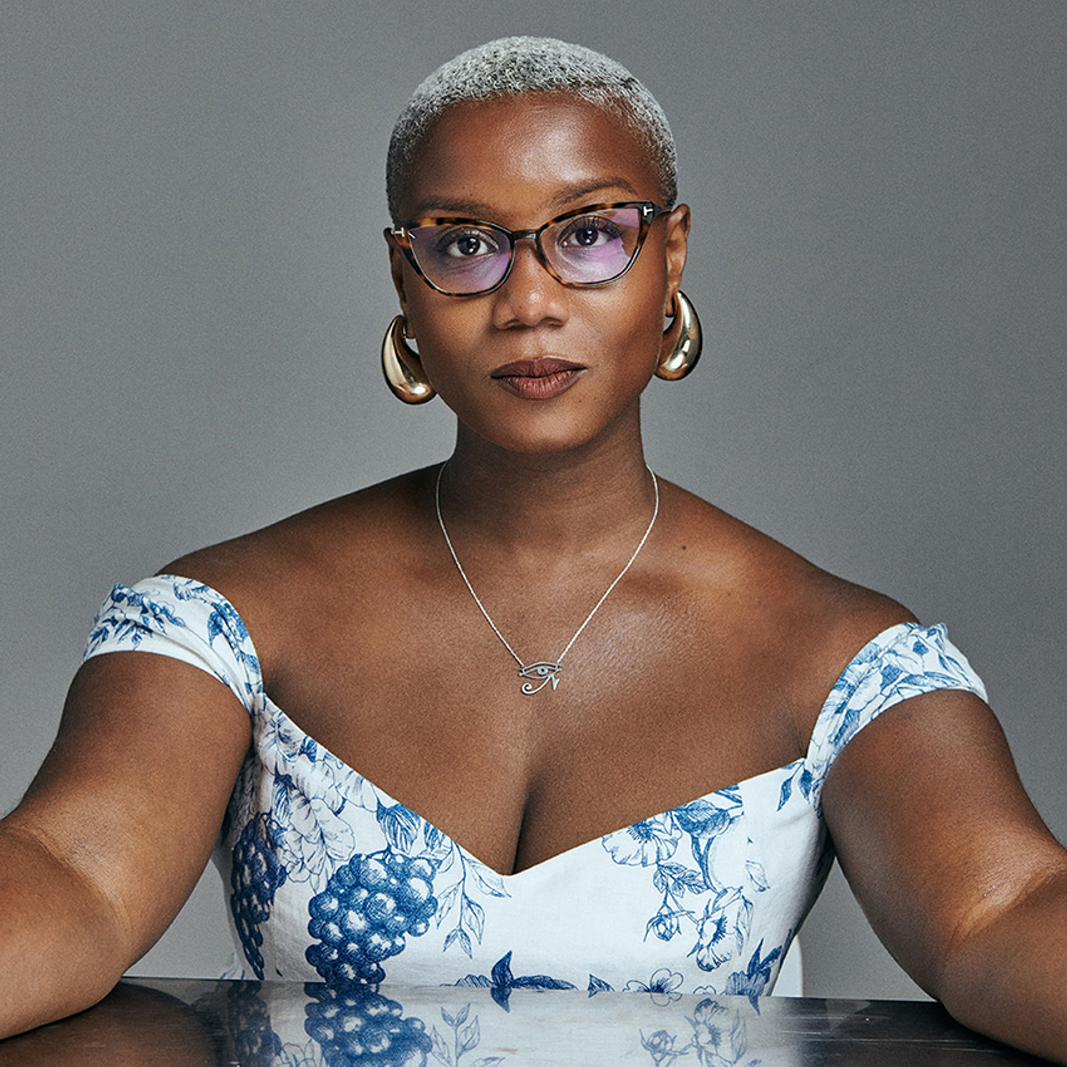 a black woman with a short buzz cut that is platinum blond looks directly into the camera. She is sitting at a reflective table and wearing a blue and white floral patterned dress, an eye of ra necklace, gold apostrophe earrings, and tortoise shell glasses.