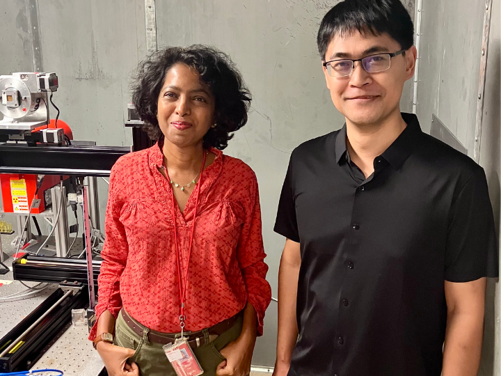 Two individuals standing in a laboratory environment, with equipment visible in the background. The person on the left is wearing a red blouse, and the person on the right is dressed in a black shirt. Both are smiling at the camera.